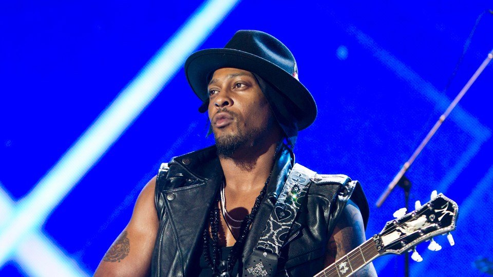 D’Angelo performing live on stage wearing a black hat and leather vest, playing guitar under blue stage lights.