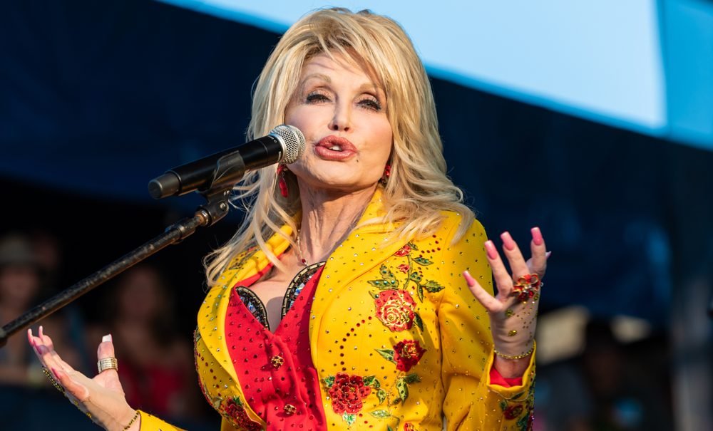 Dolly Parton performs on stage in a bright yellow sequined top during a 2025 concert, radiating her trademark warmth and energy. The country legend, whose net worth is estimated at $650 million, continues to honor the memory of her late husband, Carl Dean, through her music and philanthropy.